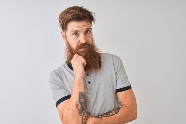 Young redhead irish man wearing grey polo standing over isolated white background looking confident at the camera smiling with crossed arms and hand raised on chin. Thinking positive. - Photo, Image