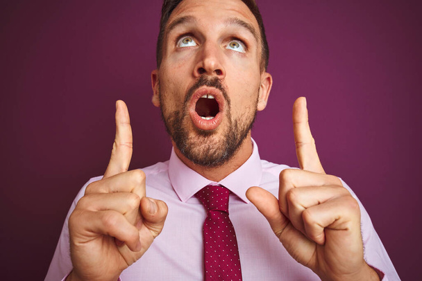 Крупный план делового человека в элегантной рубашке и галстуке на фиолетовом изолированном фоне удивлен и удивлен, глядя вверх и указывая пальцами и поднятыми руками
. - Фото, изображение