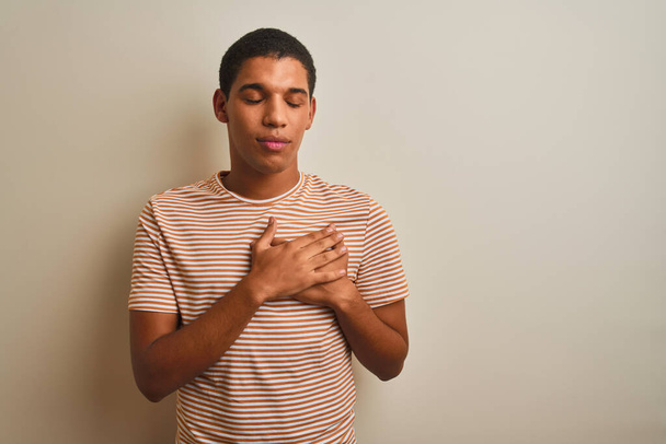 Young handsome arab man wearing striped t-shirt over isolated white background smiling with hands on chest with closed eyes and grateful gesture on face. Health concept. - Fotó, kép