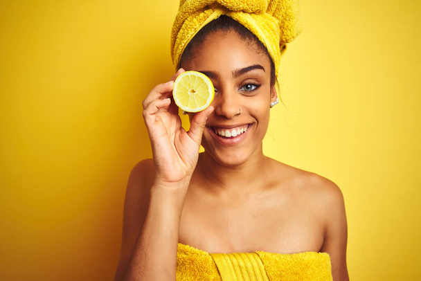 Afro woman wearing towel after shower holding slice lemon over isolated yellow background with a happy face standing and smiling with a confident smile showing teeth - Foto, afbeelding