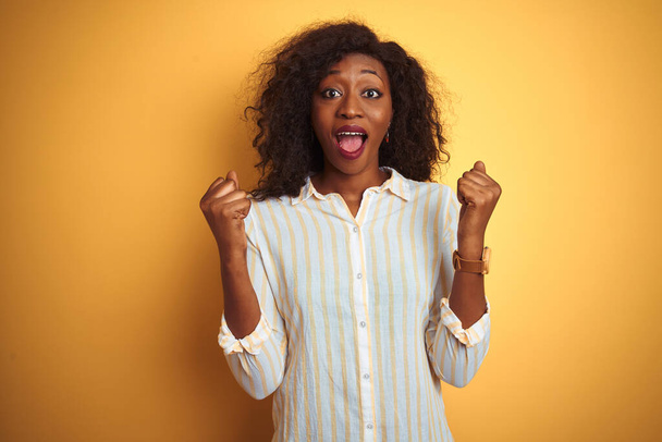 African american woman wearing striped shirt standing over isolated yellow background celebrating surprised and amazed for success with arms raised and open eyes. Winner concept. - 写真・画像
