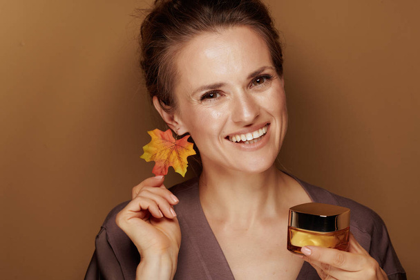 Hello autumn. Portrait of smiling young woman in a bathrobe with autumn leaf earring showing facial creme on beige background. - Fotoğraf, Görsel