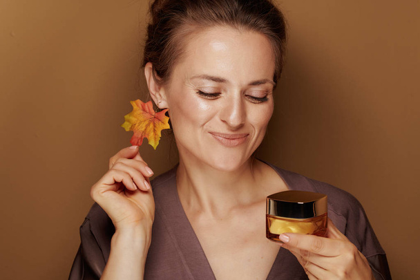 Hello autumn. Portrait of smiling modern woman in a bathrobe with autumn leaf earring looking at facial creme isolated on bronze background. - Fotoğraf, Görsel