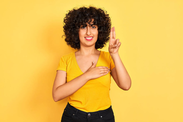 Jeune femme arabe aux cheveux bouclés portant un t-shirt debout sur un fond jaune isolé, jurant avec sourire, la main sur la poitrine et les doigts levés, prêtant serment de fidélité
 - Photo, image