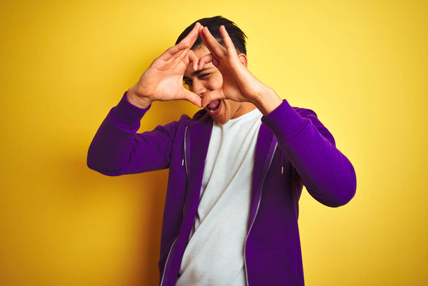 Молодой бразилец в фиолетовой толстовке стоит на изолированном желтом фоне и делает форму сердца с рукой и пальцами, улыбаясь, глядя сквозь знак
 - Фото, изображение