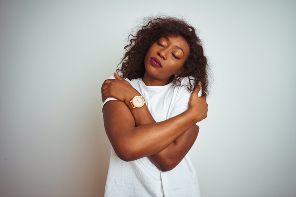 Jovem afro-americana vestindo camiseta em pé sobre fundo branco isolado Abraçando-se feliz e positivo, sorrindo confiante. Auto amor e auto cuidado
 - Foto, Imagem