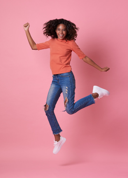 Portrait of a joyful young african woman in orange shirt jumping and celebrating over pink background. - Zdjęcie, obraz