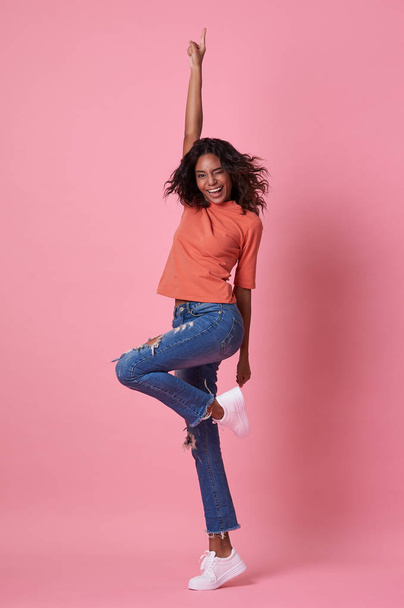 Portrait of a joyful young african woman in orange shirt jumping and celebrating over pink background. - 写真・画像