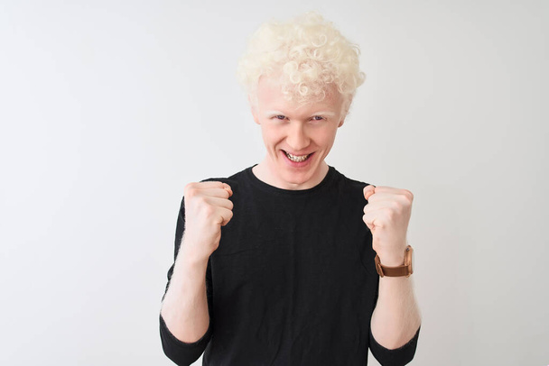 junger Albino-blonder Mann in schwarzem T-Shirt, der vor isoliertem weißen Hintergrund steht und überrascht und erstaunt über den Erfolg mit erhobenen Armen und offenen Augen feiert. Siegerkonzept. - Foto, Bild