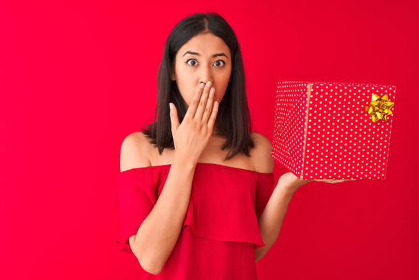 Young beautiful chinese woman holding birthday gift standing over isolated red background cover mouth with hand shocked with shame for mistake, expression of fear, scared in silence, secret concept - Φωτογραφία, εικόνα