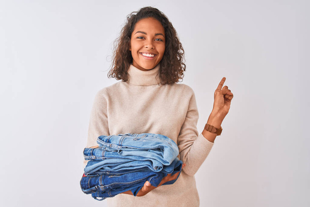 Young brazilian shopkeeper woman holding pile of jeans over isolated white background very happy pointing with hand and finger to the side - Zdjęcie, obraz