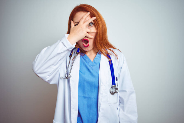 Молодая рыжеволосая женщина-врач, используя стетоскоп на белом изолированном фоне, подглядывая в ударные закрывающие лицо и глаза рукой, смотрит сквозь пальцы со смущенным выражением лица
. - Фото, изображение