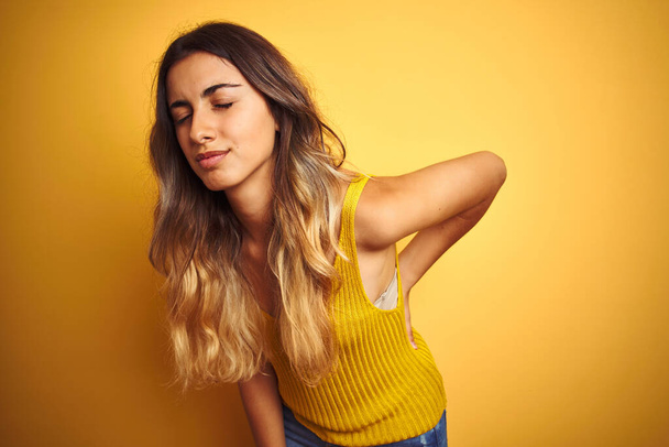 Young beautiful woman wearing t-shirt over yellow isolated background Suffering of backache, touching back with hand, muscular pain - Photo, Image