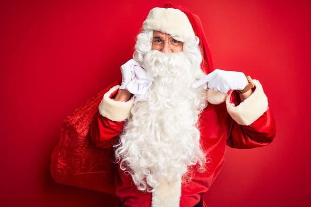 Middle age man wearing Santa costume holding sack with gifts over isolated red background with surprise face pointing finger to himself - Fotoğraf, Görsel