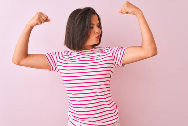 Young beautiful woman wearing striped t-shirt standing over isolated pink background showing arms muscles smiling proud. Fitness concept. - Fotó, kép