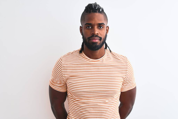 Африканский американец с косичками в полосатой футболке на изолированном белом фоне с серьезным выражением лица. Простой и естественный взгляд на камеру
. - Фото, изображение