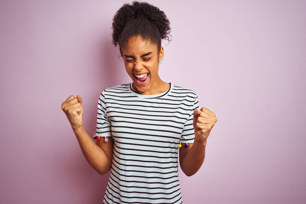 African american woman wearing navy striped t-shirt standing over isolated pink background very happy and excited doing winner gesture with arms raised, smiling and screaming for success. Celebration concept. - 写真・画像