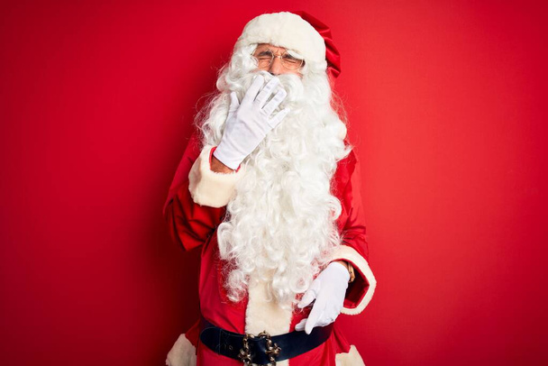 Hombre guapo de mediana edad con traje de Santa de pie sobre fondo rojo aislado aburrido bostezo cansado cubriendo la boca con la mano. Inquietud y somnolencia
. - Foto, Imagen