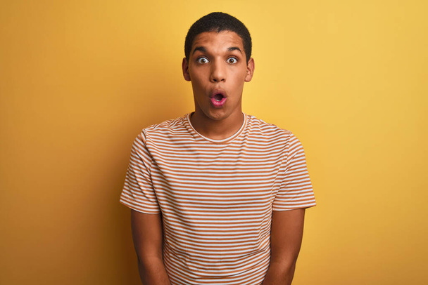 Fiatal jóképű arab férfi csíkos pólóban áll izolált sárga háttér fél, és sokkolt a meglepetés kifejezés, félelem és izgatott arc. - Fotó, kép