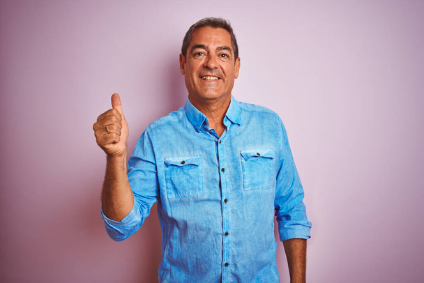 Красивый мужчина средних лет в синей джинсовой рубашке стоит на изолированном розовом фоне и делает счастливый жест вверх рукой. Утверждение выражения лица, смотрящего в камеру с успехом
. - Фото, изображение