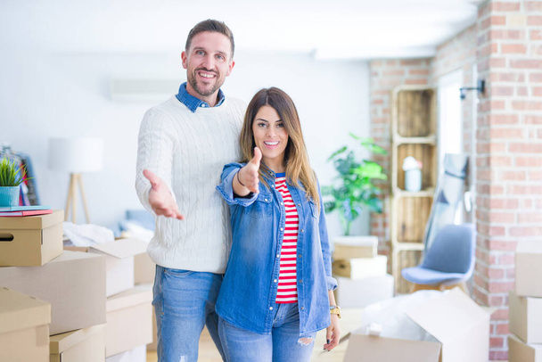 Young beautiful couple standing at new home around cardboard boxes smiling friendly offering handshake as greeting and welcoming. Successful business. - Photo, Image