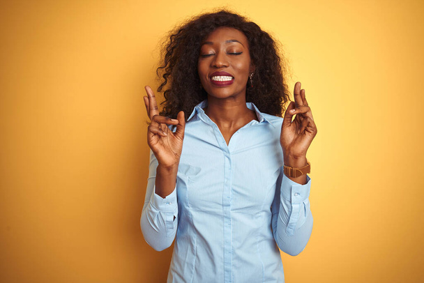 Afro-Amerikaanse zakenvrouw draagt elegant shirt over geïsoleerde gele achtergrond gebaren vinger gekruist glimlachend met hoop en ogen dicht. Geluk en bijgelovig concept. - Foto, afbeelding