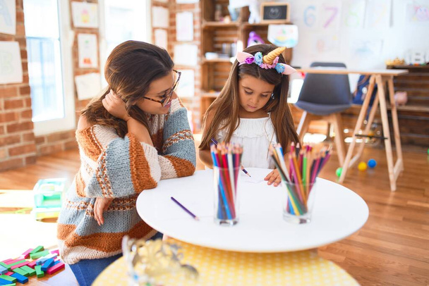Bela professora e criança vestindo unicórnio desenho diadema usando papel e lápis em torno de lotes de brinquedos no jardim de infância
 - Foto, Imagem