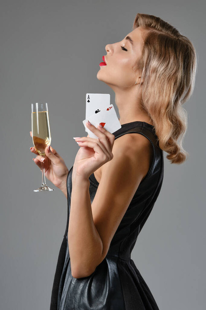 Menina loira em vestido preto segurando duas cartas de jogo e vidro de champanhe, posando contra fundo cinza. Jogos de azar, poker, cassino. Close-up. - Foto, Imagem