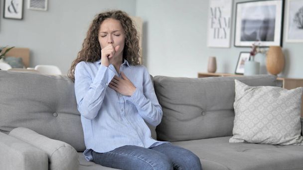 Больная молодая кудрявая женщина кашляет, сидя на диване
 - Фото, изображение