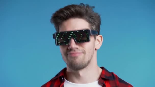 Hipster man in glowing neon glasses moving to the music rhythm on blue studio background. Dancing positive comic guy. 4k - Metraje, vídeo