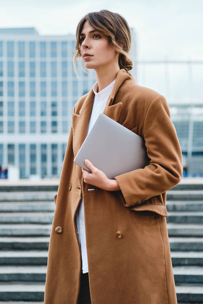 Fiatal vonzó elegáns üzletasszony kabátban laptoppal magabiztosan néz félre a város utcáján - Fotó, kép