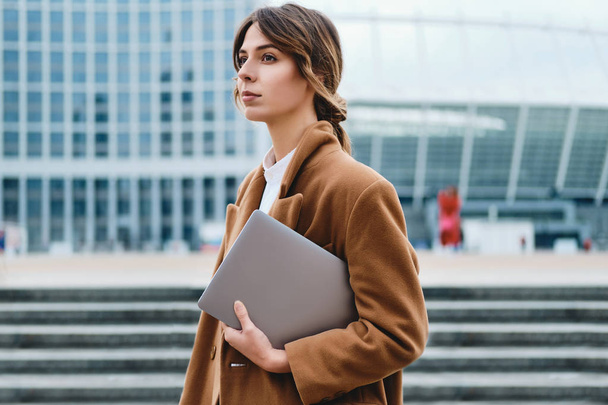 Jeune belle femme d'affaires en manteau avec ordinateur portable regardant attentivement loin sur la rue de la ville
 - Photo, image