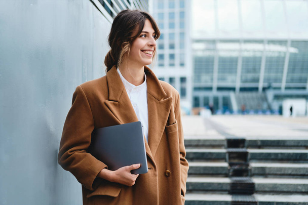 junge, gut gelaunte Geschäftsfrau im Mantel mit Laptop schaut fröhlich auf der Straße der Stadt weg - Foto, Bild