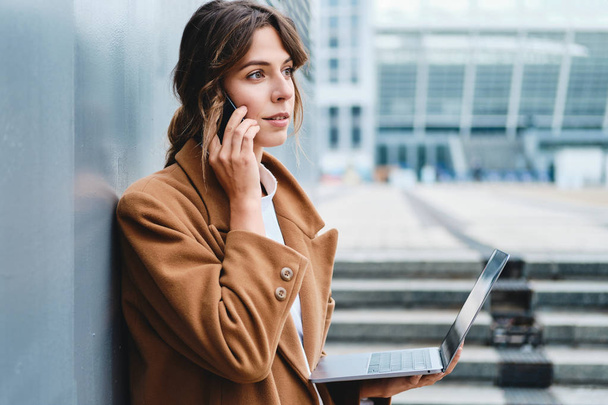 Jovem mulher de negócios elegante no casaco confiantemente falando no celular trabalhando no laptop na rua da cidade
 - Foto, Imagem