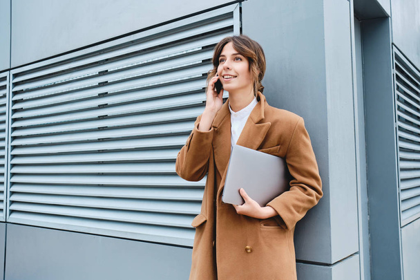 Joven mujer de negocios bonita en abrigo con portátil felizmente hablando en el teléfono celular al aire libre
 - Foto, Imagen