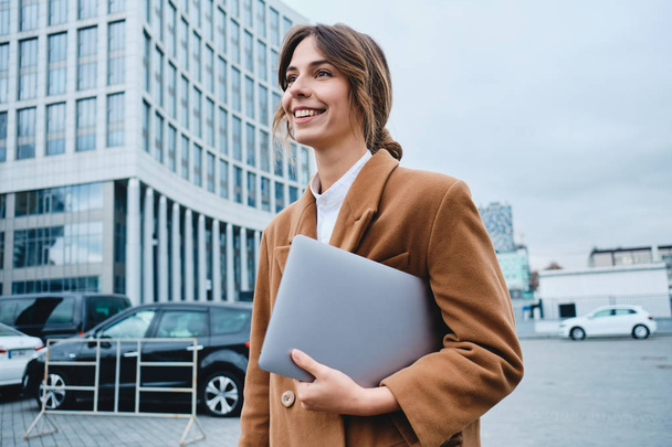 Jeune femme d'affaires souriante attrayante en manteau avec ordinateur portable regardant joyeusement loin en marchant dans la rue de la ville
 - Photo, image
