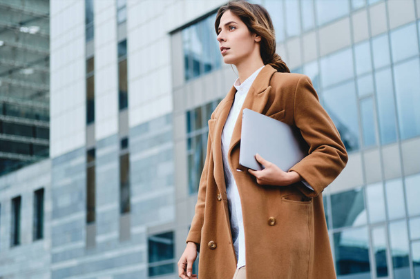Young serious businesswoman in coat with laptop thoughtfully walking through city street - Φωτογραφία, εικόνα