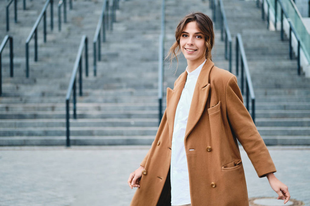 Young attractive stylish woman in coat happily looking in camera outdoor - Fotografie, Obrázek