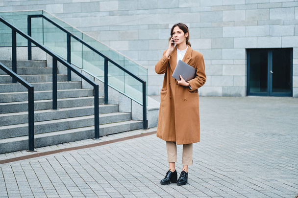Young attractive businesswoman in coat with laptop confidently talking on cellphone on city street - Foto, Imagem