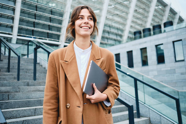 junge hübsche Geschäftsfrau im Mantel mit Laptop, die freudig nach draußen schaut - Foto, Bild