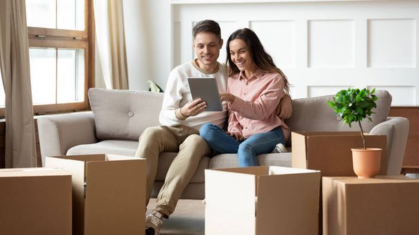 Happy couple using digital tablet sit on sofa with boxes - Фото, зображення