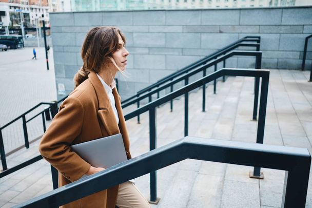 Boční pohled na mladé atraktivní podnikatelky v kabátě s notebookem sebevědomě chůze na ulici - Fotografie, Obrázek