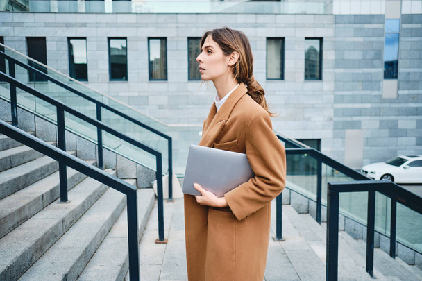 Vista lateral da jovem mulher de negócios lindo no casaco com laptop confiantemente olhando para o lado ao ar livre
 - Foto, Imagem