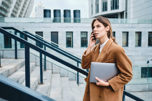 Widok z boku młody poważny businesswoman w płaszczu z laptopem rozmawia przez komórkę idzie po schodach na zewnątrz - Zdjęcie, obraz