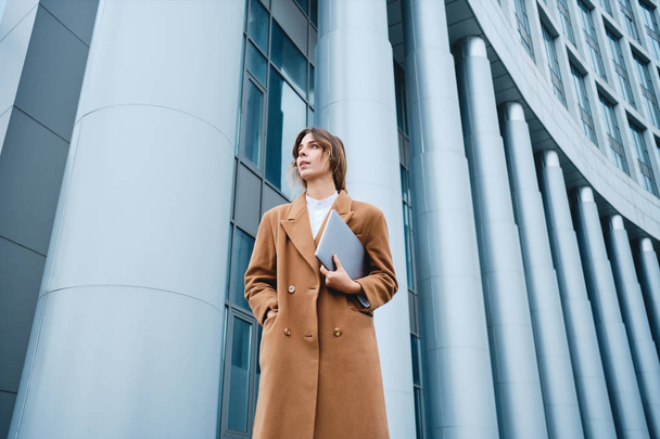 Joven mujer de negocios con estilo y confianza en el abrigo con portátil mirando atentamente al aire libre
 - Foto, Imagen