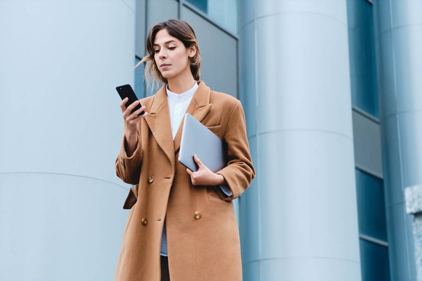 Jeune femme d'affaires décontractée confiante en manteau avec ordinateur portable en utilisant un téléphone portable extérieur
 - Photo, image