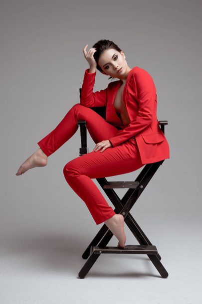 молодая красивая женщина сидит на режиссерском стуле. модель брюнетки в красном костюме на белом фоне
. - Фото, изображение
