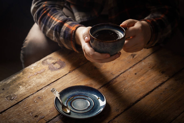 Gros plan noir americano café en tasse bleue à la main masculine, soucoupe avec cuillère à thé vintage sur fond marron table de village vintage en bois. Concept matin petit déjeuner homme seul, célibataire
 - Photo, image