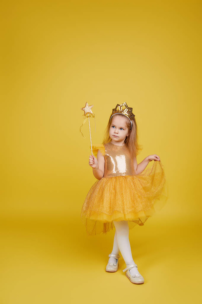 Joyful little girl with long hair in a tulle golden dress and princess crown holding a magic wand  on yellow background. Celebrating a colorful carnival for kids, expressing positive birthday - Photo, Image