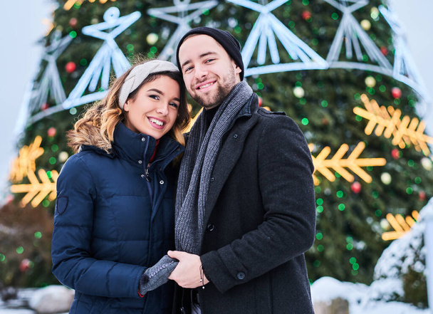 Happy couple wearing warm clothes hold hands and look at each other, standing near a city Christmas tree, enjoying spending time together. - Fotó, kép